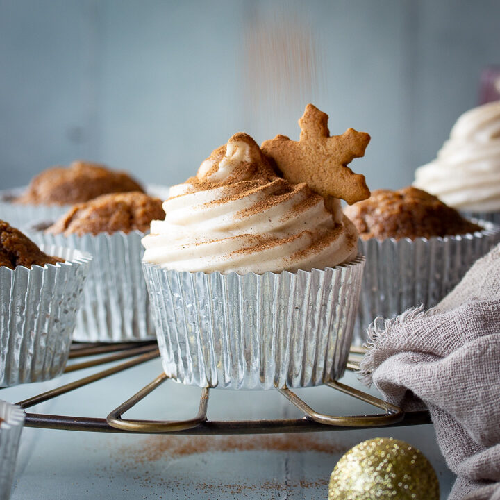 A close up of a gingerbread cupcake topped with a gingerbread biscuit snowflake on a cooling rack being sprinkled with extra cinnamon. There are more cupcakes, gold baubles and a light brown linen napkin in the background. Set on a cool grey wood effect backdrop.