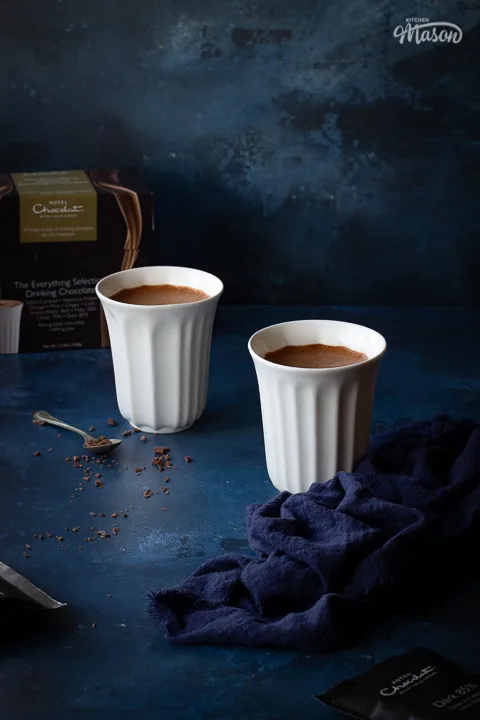 2 white pod cups filled with hot chocolate on a hand painted deep blue backdrop. There's also a box of Hotel Chocolat hot chocolate sachets, a teaspoon filled with grated chocolate and a blue linen napkin in the background.