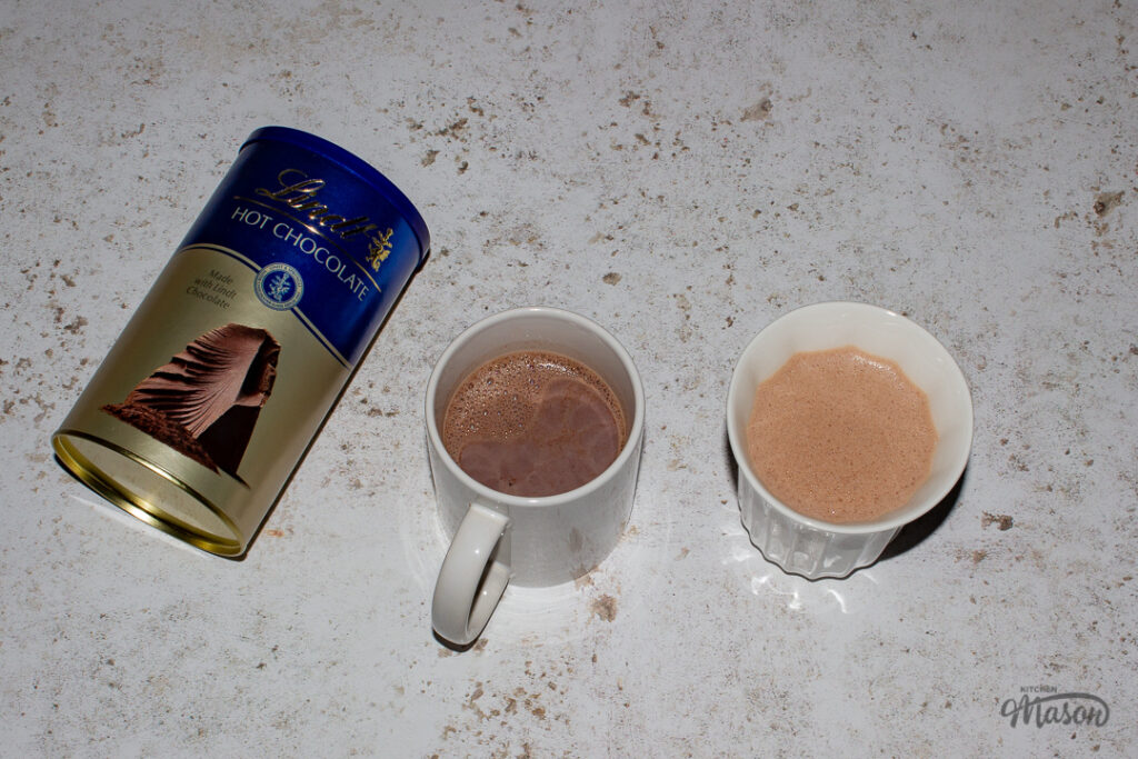 2 mugs filled with hot chocolate and a tub of Lindt hot chocolate powder against a mottled white backdrop.