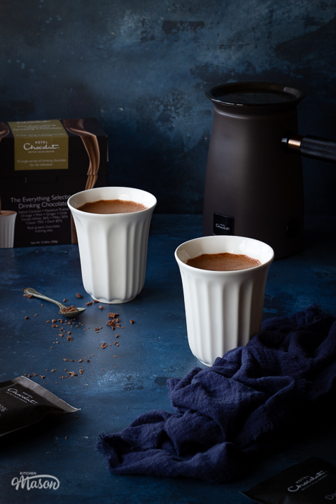 2 white pod cups filled with hot chocolate and the Hotel Chocolate velvetiser on a hand painted deep blue backdrop. There's also a box of Hotel Chocolat hot chocolate sachets, a teaspoon filled with grated chocolate and a blue linen napkin in the background.