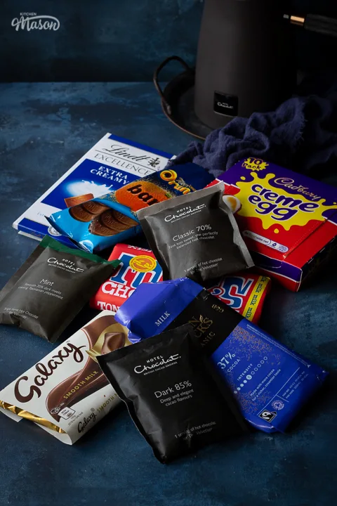 The Hotel Chocolat Velvetiser in the background of a shot filled with various chocolate bars in their wrappers. Set against a deep blue hand painted backdrop.