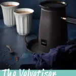 Close up of the Hotel Chocolat Velvetiser on a grey tray with a blue linen napkin on the side. There's also 2 white pod cups filled with hot chocolate and a spoon topped with grated chocolate in the background. Set against a deep blue hand painted backdrop. A text overlay says 'The Velvetiser, is it worth it?'