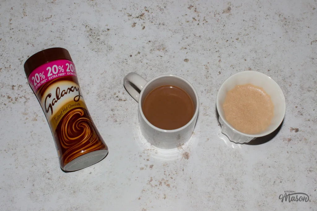 Two mugs filled with hot chocolate and a tub of Galaxy hot chocolate powder set against a mottled white backdrop.