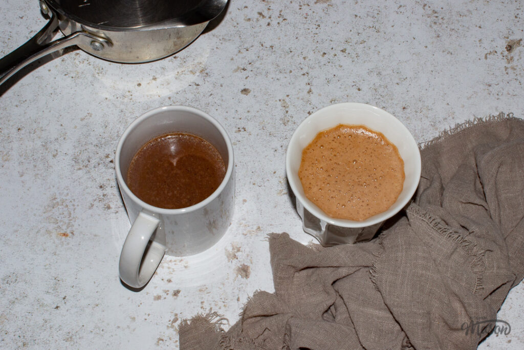 2 Mugs filled with hot chocolate set against a mottled white backdrop along with an empty saucepan and a light brown linen napkin.