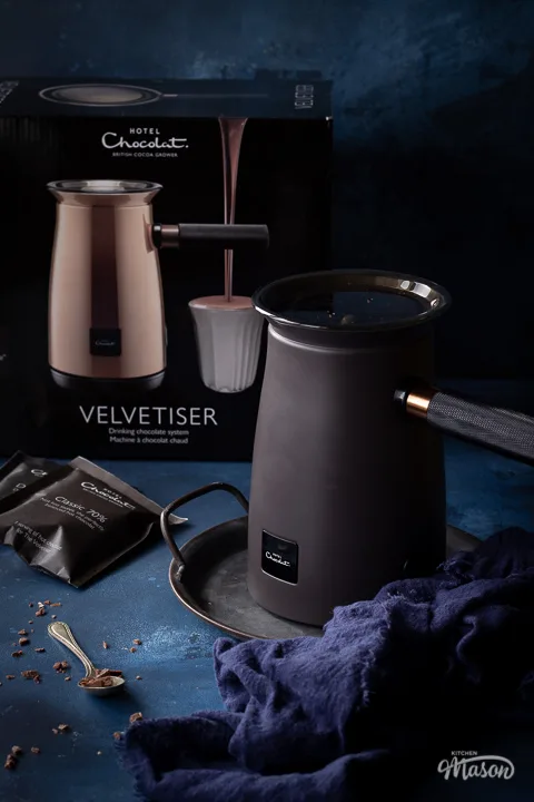 The Hotel Chocolat velvetiser on a grey tray with a blue linen napkin on the side. The velvetiser box, some chocolate sachets and a spoon with grated chocolate on are also in the background. Set against a hand painted deep blue backdrop.