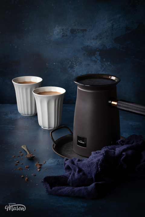The Hotel Chocolat Velvetiser on a grey tray with a blue linen napkin on the side. There's also 2 white pod cups filled with hot chocolate and a spoon topped with grated chocolate in the background. Set against a deep blue hand painted backdrop.