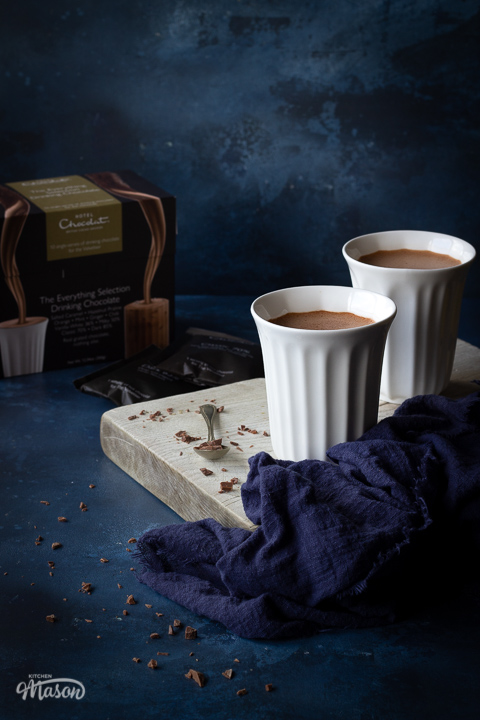 Two white pod cups filled with hot chocolate set on a white wood board with a teaspoon topped with chopped chocolate and a blue linen napkin. There's a box of Hotel Chocolat hot chocolate sachets in the background and it's all set against a deep blue hand painted backdrop.