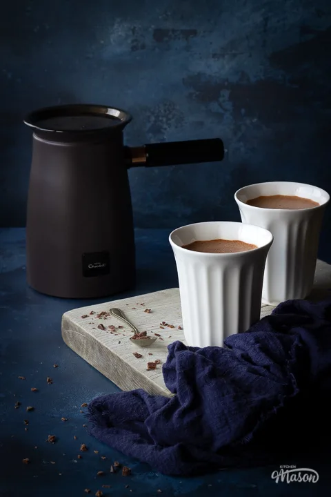 2 white pod cups filled with hot chocolate on a white wood board with a spoon and some grated chocolate on the side. The Hotel Chocolat velvetiser machine is in the background along with a blue linen napkin. Set on a deep blue hand painted backdrop.