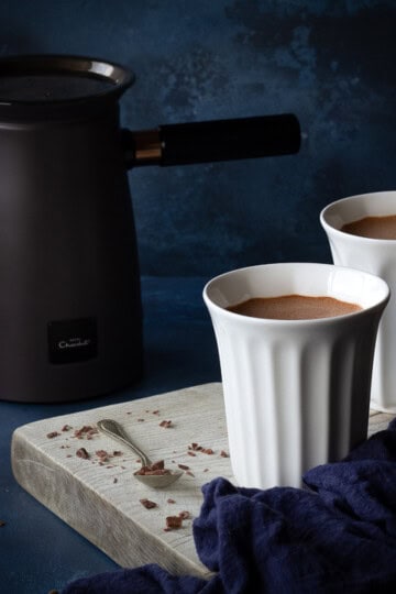 2 white pod cups filled with hot chocolate on a white wood board with a spoon and some grated chocolate on the side. The Hotel Chocolat velvetiser machine is in the background along with a blue linen napkin. Set on a deep blue hand painted backdrop.