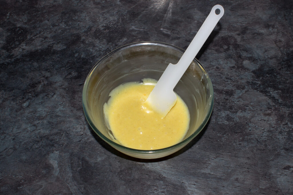 A glass bowl with melted white chocolate and orange juice inside on a kitchen worktop.