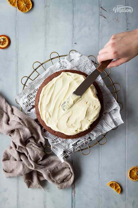 A chocolate orange cake on a cake rack topped with baking paper, having white chocolate filling spread on top with a pallet knife. Set on a cool grey painted wood effect backdrop, there's also orange segments and a light brown linen napkin in the background.