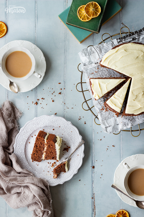 Top down view of a sliced chocolate orange cake on a wire rack, two cups of tea and a partially eaten slice of cake on a white plate with a fork. Set against a cool grey hand painted wood effect backdrop, there are also some books, dried orange segments and a light brown linen napkin in the background.