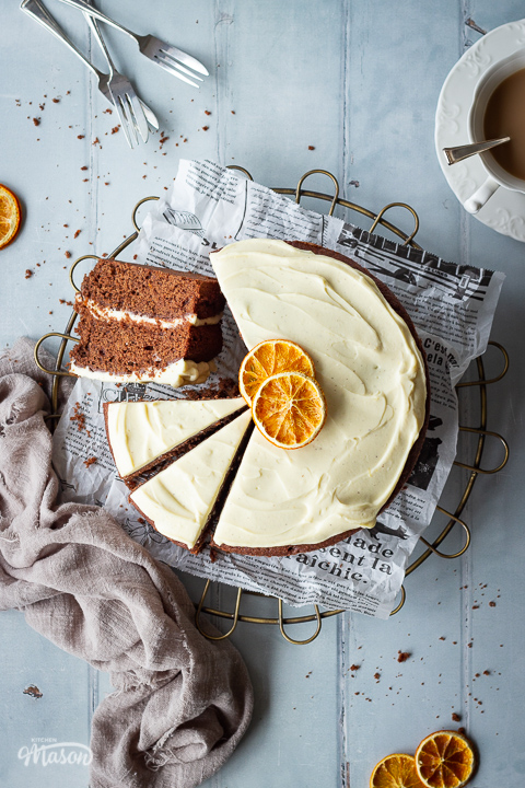A close up top down view of a sliced chocolate orange cake on a round wire rack. One of the slices is turned on it's side so you can see inside. Set on a cool grey hand painted wood effect backdrop, there is also a cup of tea, some small forks, dried orange segments, a light brown linen napkin and some cake crumbs in the background.