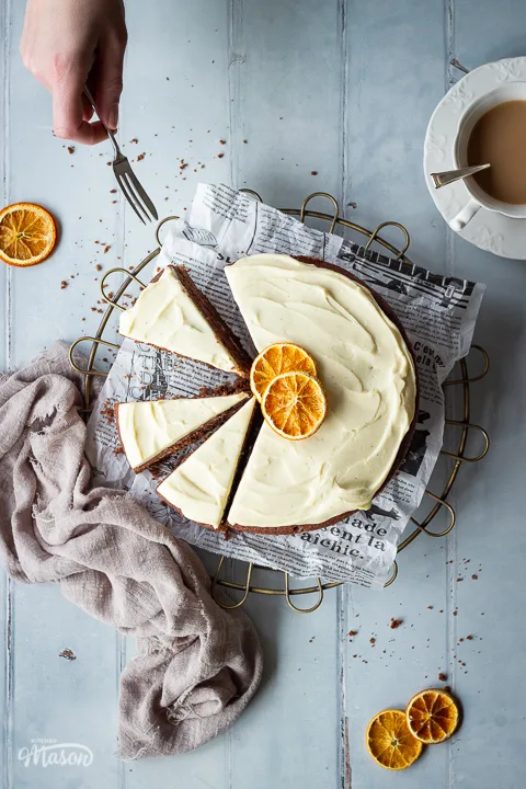 A hand reaching in with a small fork to a sliced chocolate cake set on a round wire stand. Set on a cool grey hand painted wood effect backdrop, there is also a cup of tea, some dried orange segments, a light brown linen napkin and some cake crumbs in the background.