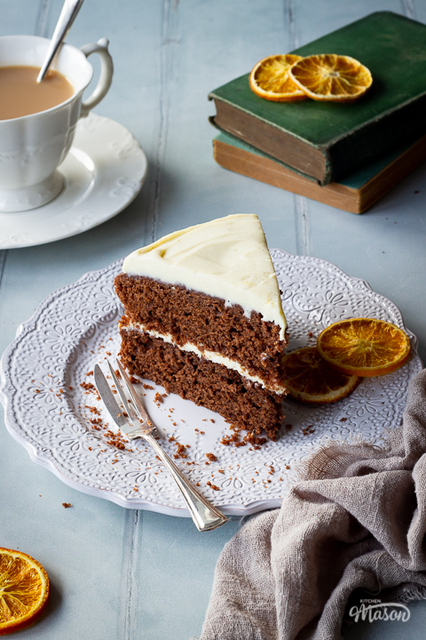 Angled front view of a slice of chocolate orange cake on a white patterned plate with a small fork and 2 dried chocolate orange segments on the side. Set against a cool grey hand painted ood effect backdrop, there is also a cup of tea, two books, a light brown linen napkin and some dried orange segments in the background.