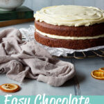 Front view of a whole chocolate orange cake on a round wire cake stand. Set on a cool grey painted wood effect backdrop, there is also a bowl of white chocolate filling, two books, dried orange segments and a light brown linen napkin in the background. A text overlay says "Easy Chocolate Orange Cake".