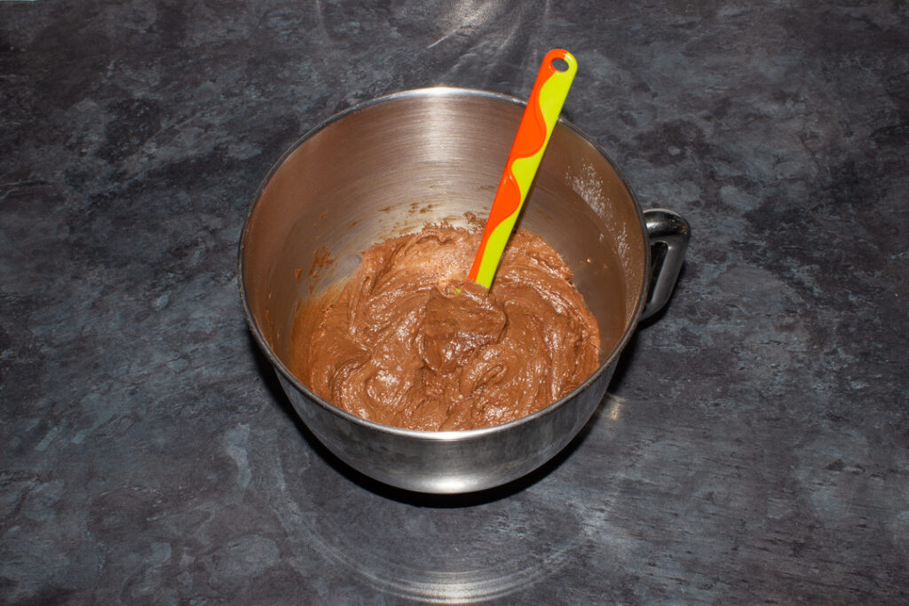 Chocolate orange cake batter in the bowl of an electric stand mixer with a green spatula on a kitchen worktop.