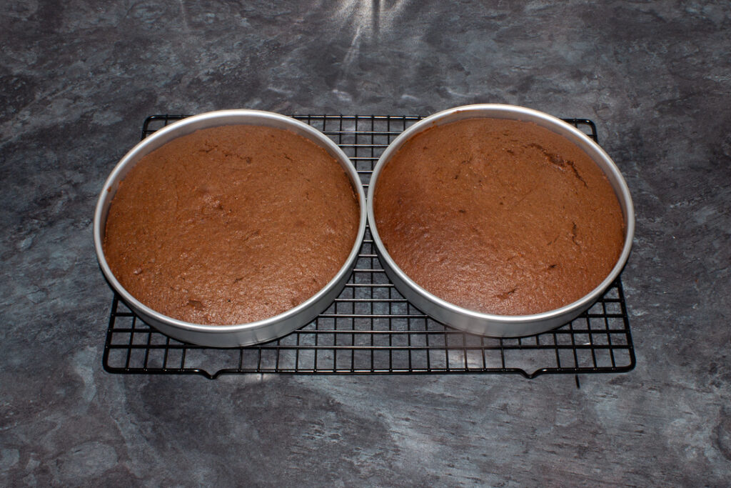 Two baked chocolate orange cakes in lined circular tins on a cooling rack set on a kitchen worktop.
