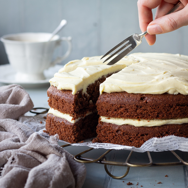 Someone reaching in to a chocolate orange cake with a fork. Set against a cool grey wood effect backdrop, there's also a cup of tea and a light brown linen napkin scrunched up in the background.