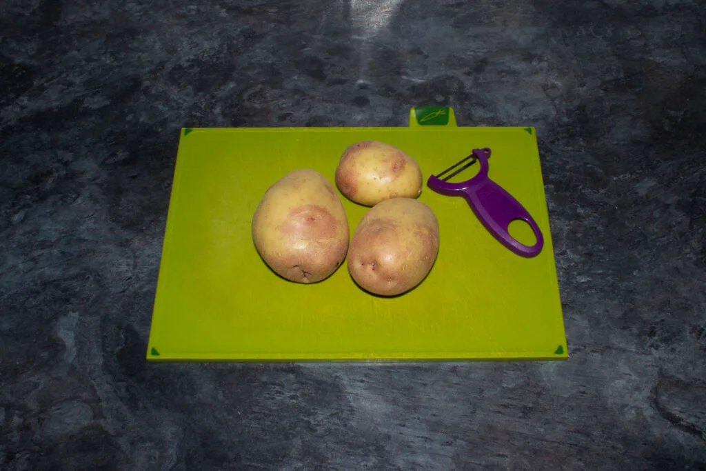 3 potatoes on a green chopping board with a purple potato peeler. Set on a kitchen worktop.