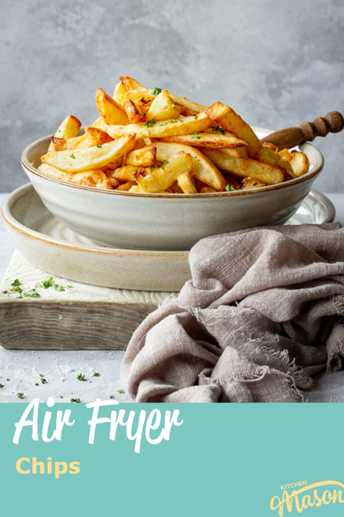 Front view of a fork in a bowl of air fryer chips set on a plate over a white board. Set on a grey painted plaster backdrop, there is also some scattered chopped parsley and a light brown linen napkin in the background. A text overlay says "air fryer chips".