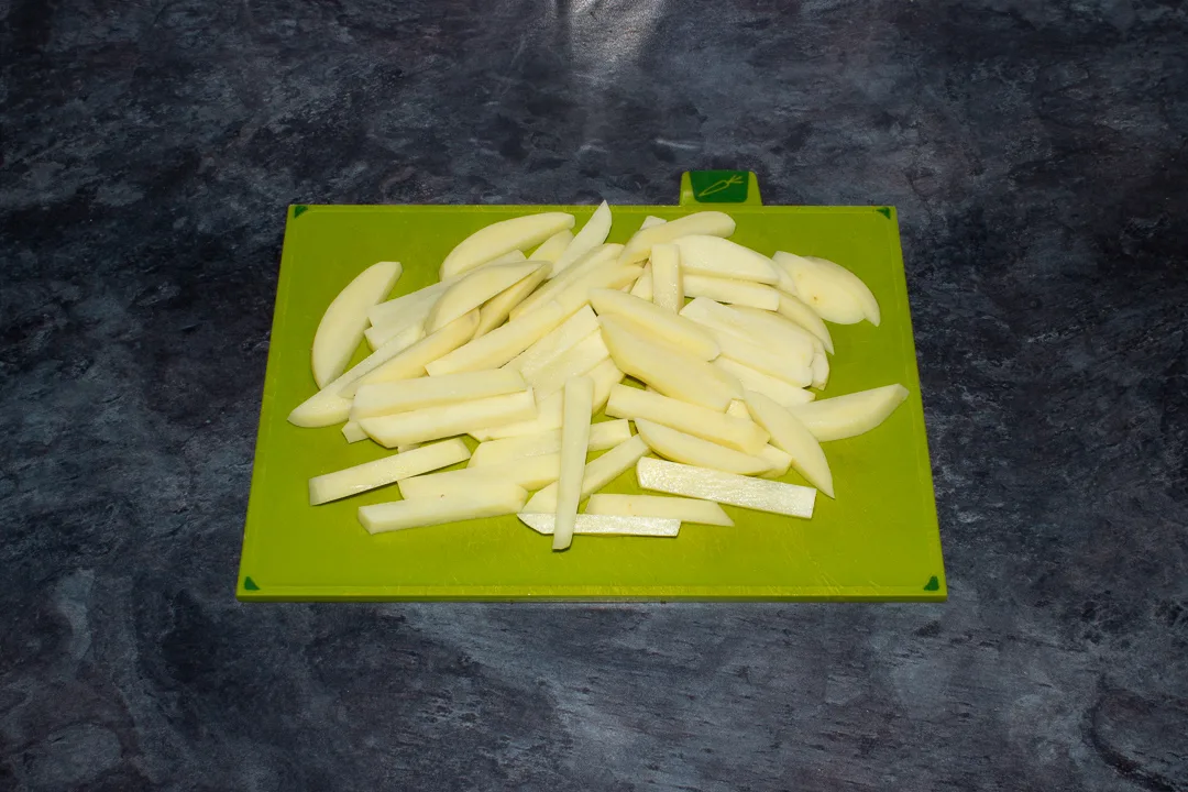 Peeled and sliced potatoes on a green chopping board set on a kitchen worktop.