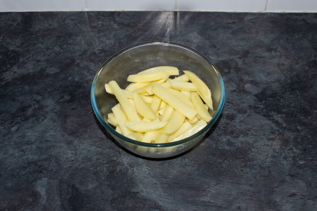 Uncooked chips coated in olive oil and salt in a large glass bowl on a kitchen worktop.