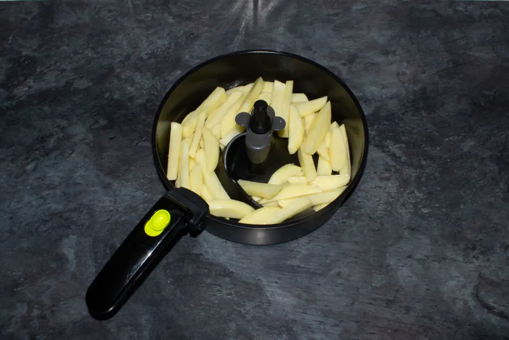 Uncooked chips coated in olive oil and salt in an air fryer pan set on a kitchen worktop.