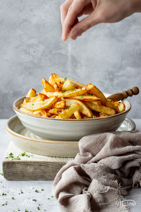 Front view of a hand sprinkling salt into a bowl of air fryer chips, set on a plate over a white board. Set on a grey painted plaster backdrop, there is also some scattered chopped parsley and a light brown linen napkin in the background.