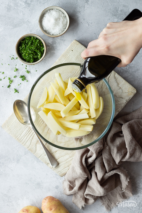 Olive oil being poured over uncooked chips in a glass bowl set on a white wood chopping board with a serving spoon. Set over a grey painted plaster backdrop, there is also a pot of chopped parsley, a pot of sea salt, 2 potatoes and a light brown linen napkin in the background.