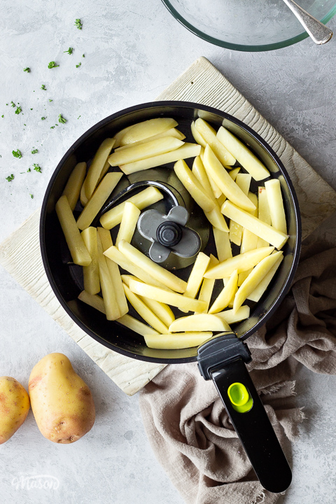 Uncooked chips coated in olive oil and sea salt in an air fryer pan set on a white wood board. There is also an empty glass bowl with a serving spoon inside, 2 potatoes, a light brown linen napkin and a scattering of chopped parsley, all set over a grey painted plaster backdrop.