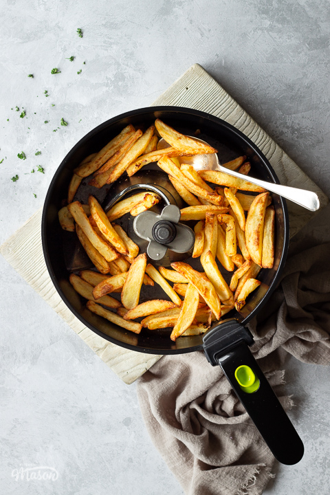 Cooked chips and a serving spoon in an air fryer pan set on a white wood board. There is a scattering of chopped parsley and a light brown linen napkin in the background. The shot is set on a grey painted plaster backdrop.