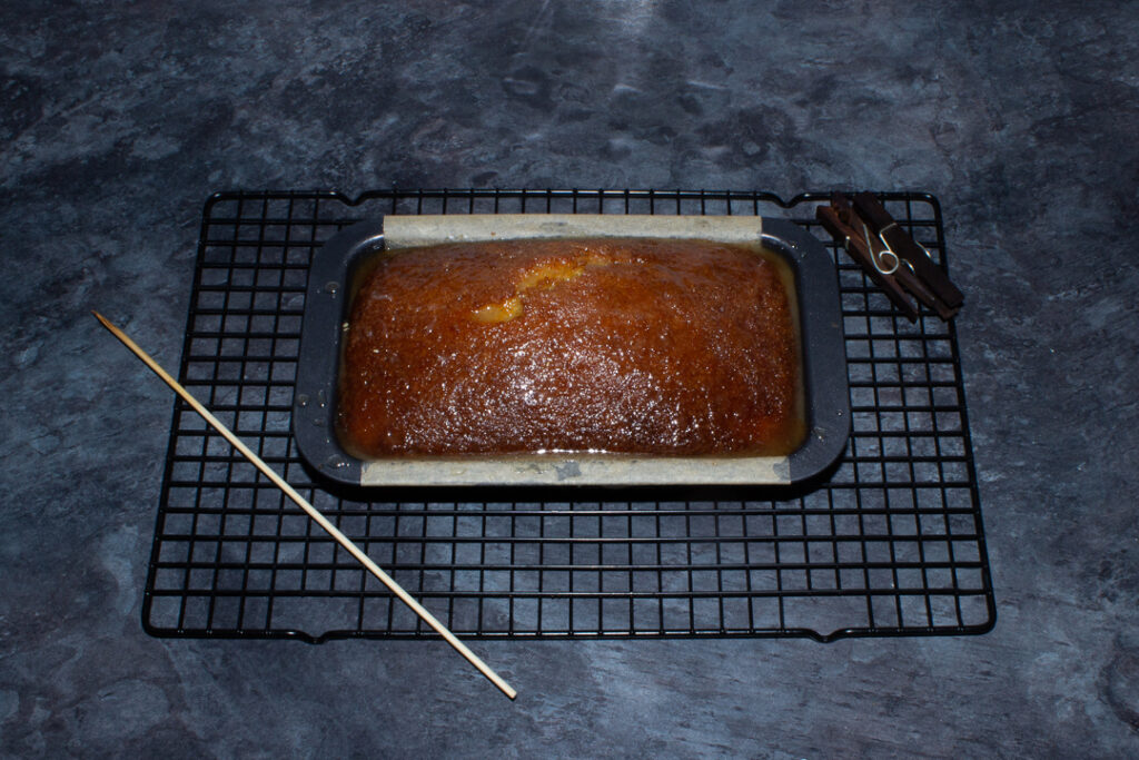 A lemon drizzle cake that's just had the drizzle poured over the top, set on a wire rack on a kitchen worktop.