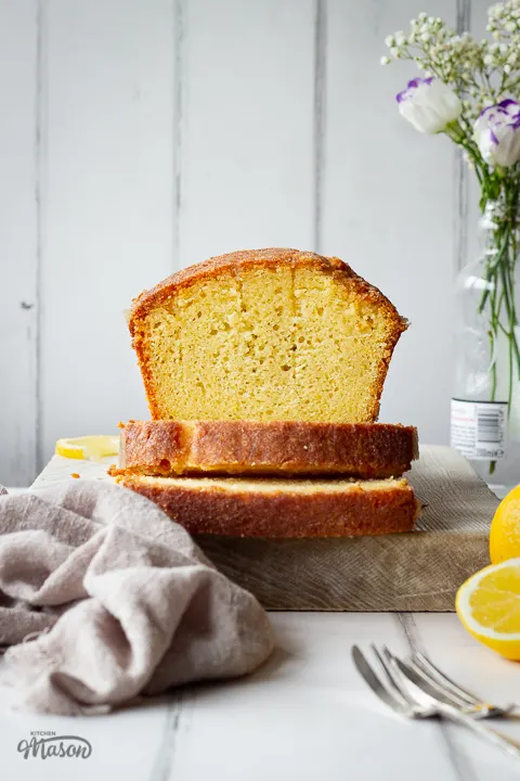 A front on view of a sliced lemon drizzle cake on a white wood board set against a white painted wood effect backdrop. There is a small glass bottle filled with white flowers off to the side, a half lemon, two forks and a light brown linen napkin in the background.