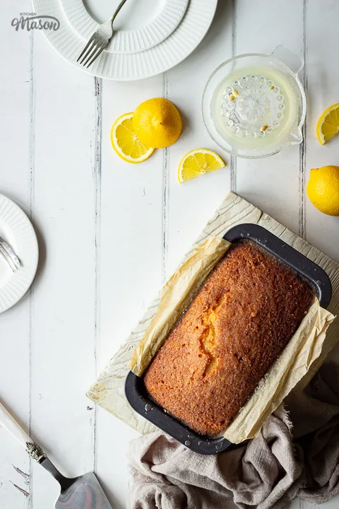 A moist lemon drizzle cake in a loaf pan on a white wooden board set against a white painted wood effect backdrop. There's also white plates, small forks, lemon slices, a lemon juicer, a cake slice and a light brown linen napkin in the background.