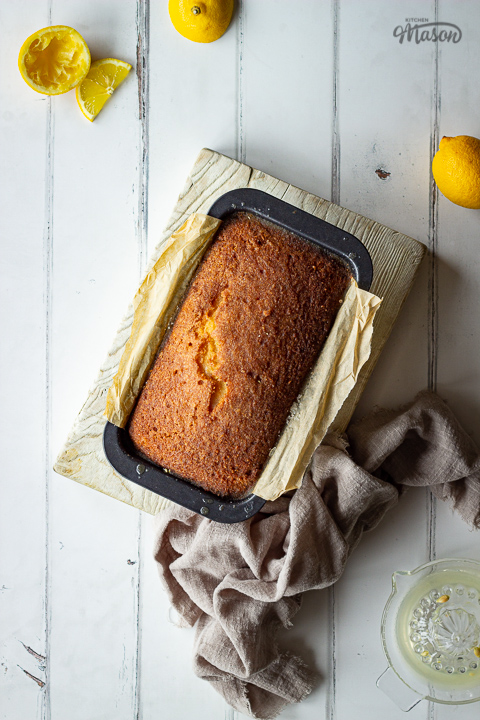 A whole lemon drizzle loaf cake in a lined pan on a white wood board. Set on a white painted wood effect backdrop, there are also lemon slices, half lemons, a light brown linen napkin and a lemon juicer in the background.