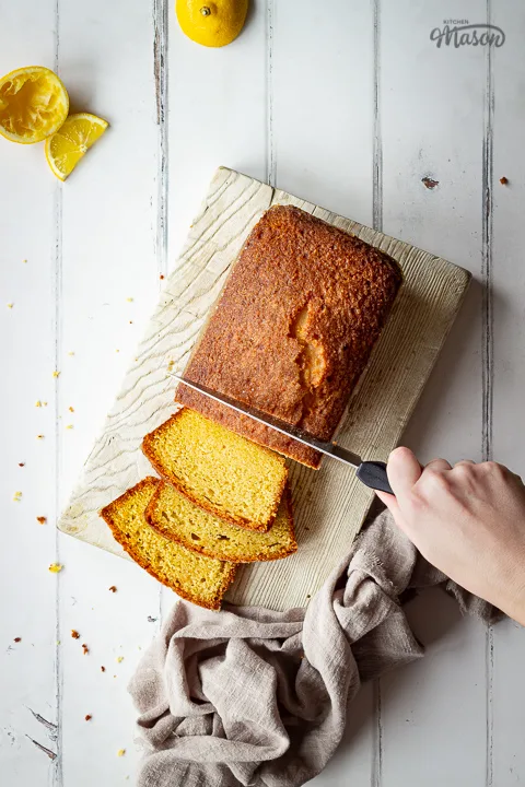 A top down view of someone slicing a moist lemon drizzle cake with a knife on a white wood board. Set on a white wood effect backdrop, there are also lemon slices, squeezed lemon halves and a light brown linen napkin in the background.