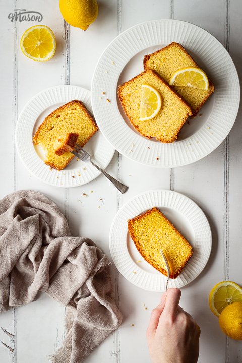 3 White plates topped with slices of moist lemon drizzle cake in a triangle shape, and a hand reaching into the shot with a fork. There are also lemon slices, squeezed lemon halves and a light brown linen napkin in the background and it's all set on a white painted wood effect backdrop.