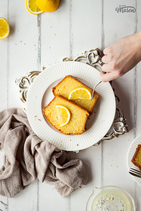A white plate set on an ornate white wood board, topped with 2 slices of moist lemon drizzle cake, lemon slices and a hand reaching into the shot with a fork. All set on a white painted wood effect backdrop, there are also lemon slices, squeezed lemon halves, a lemon juicer, a light brown linen napkin and a small plate with another slice of lemon drizzle cake in the background.