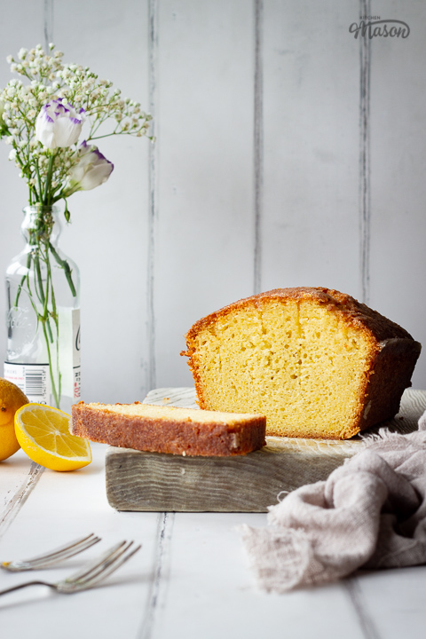 A front on view of a sliced lemon drizzle cake on a white wood board set against a white painted wood effect backdrop. There is a small glass bottle filled with white flowers, a whole lemon, two forks and a light brown linen napkin in the background.