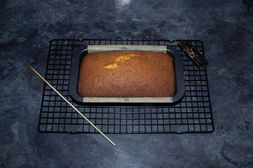 A lemon drizzle loaf cake on a wire rack with a skewer. The cake has been poked all over to make holes for the drizzle.
