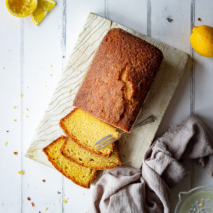 A square top down view of a half sliced moist lemon drizzle cake set on a white wood board with a fork. Set on a white wood effect backdrop, there are also lemon slices, squeezed lemon halves, a lemon juicer and a light brown linen napkin in the background.