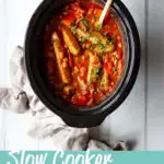 A top down view of a slow cooker filled with sausage casserole with beans. Set against a white wood effect backdrop there is a wooden spoon in the bowl and a light brown linen napkin to the side. A text overlay says "slow cooker sausage casserole"