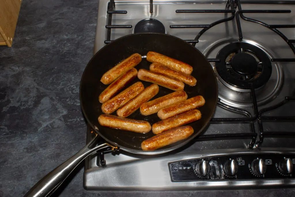 12 browned sausages frying in a pan on a stove.