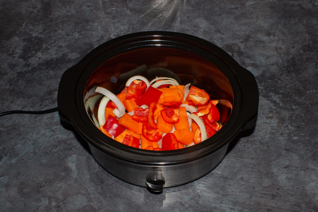 Chopped onion, carrots and pepper in a slow cooker bowl on a kitchen worktop.