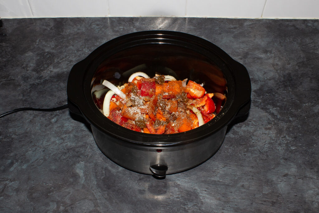 Chopped onion, carrots, pepper, spices and seasoning in a slow cooker bowl on a kitchen worktop.