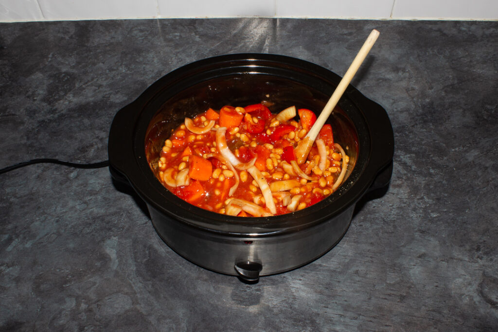 Chopped vegetables, spices, tomatoes and beans in a slow cooker bowl with a wooden spoon inside on a kitchen worktop.