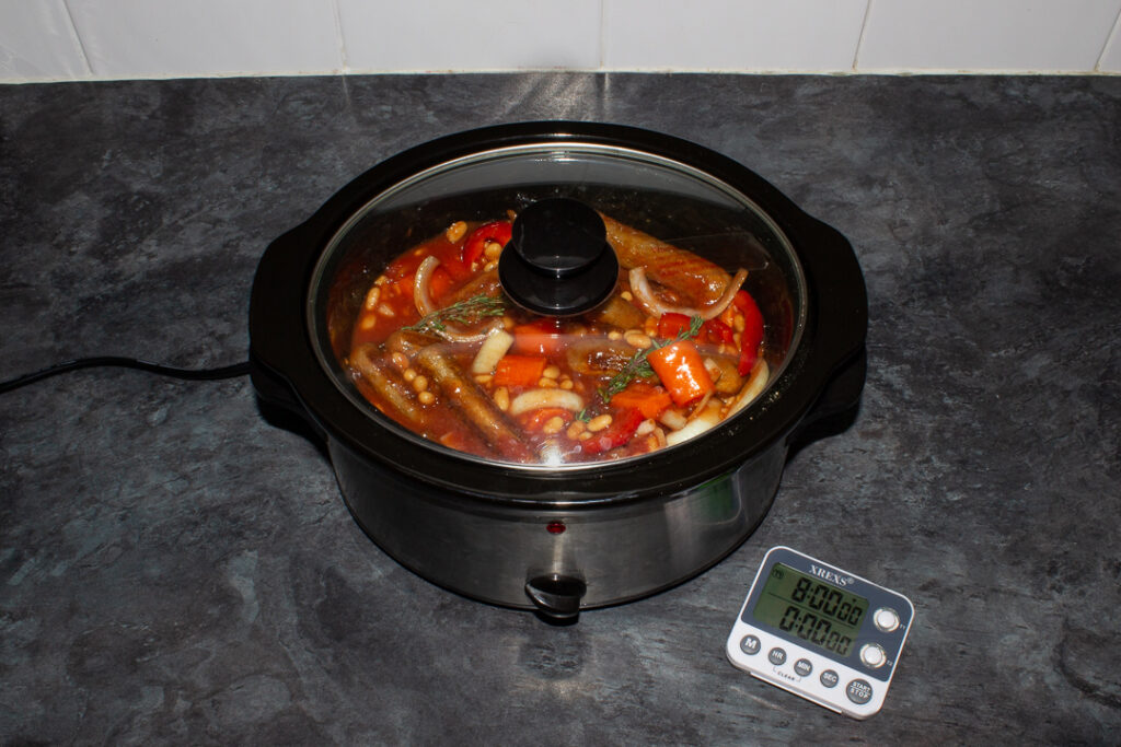 Slow cooker sausage casserole with beans in a slow cooker on a kitchen worktop with a timer next to it saying '8 hours'.