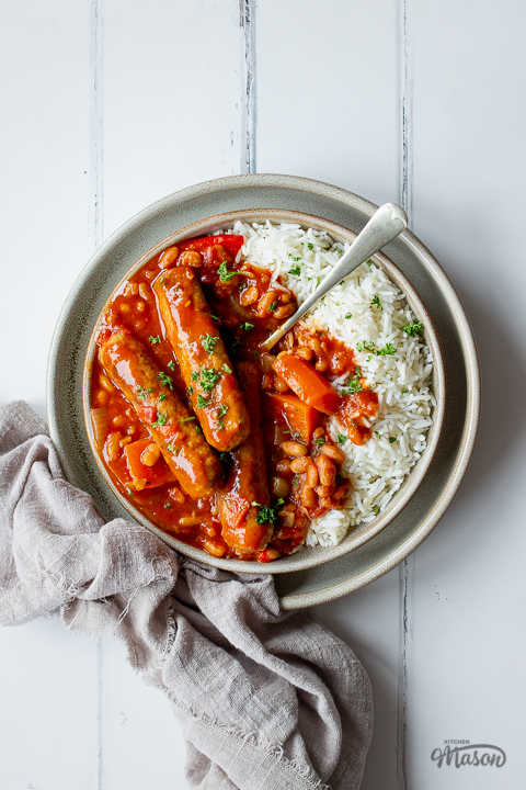 A close up top down view of slow cooker sausage casserole with beans and rice on a small plate set over a larger plate. There's a fork in the casserole, a light brown linen napkin to the side and it's set over a white wood effect backdrop.