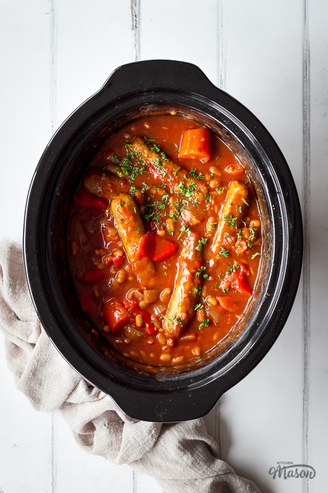 A close up top down view of a slow cooker sausage casserole in a slow cooker on a white wood effect backdrop with a light brown linen napkin to the side.